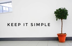 Photo of white wall. Small tree in a pot on the right and the words 'KEEP IT SIMPLE' on the wall.